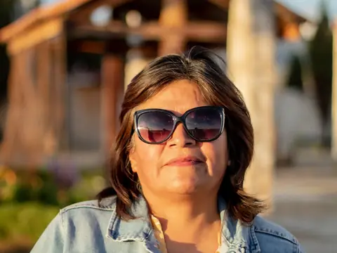 A woman in a denim jacket and sunglasses outside a wooden building on a sunny day